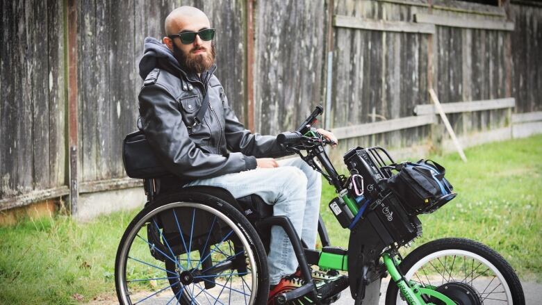 A bald man with sunglasses sits on a wheelchair with a bike-like extension. He is in a garden.