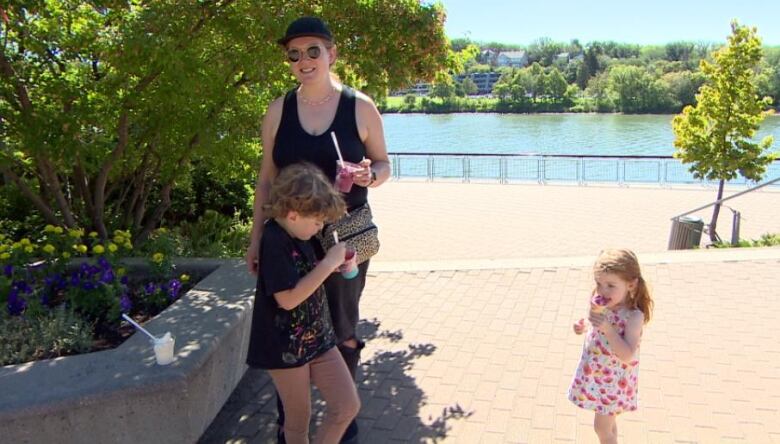 A woman stands outside near a river with two young children on a sunny day.