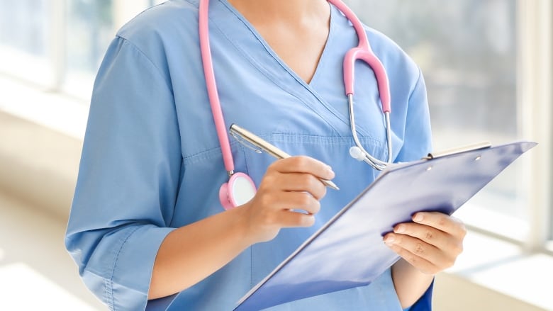 Young African-American nurse wearing blue scrubs, with stethoscope around her neck, holds clipboard in left hand and pen in right hand.