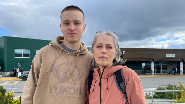 Two volunteers stand in front of the Whitehorse airport. They are wearing pastel colours and looking pensively at the camera.