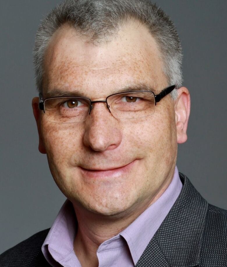 A man wearing  a purple shirt and suit jacket looks into the camera while wearing glasses.