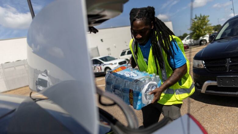 A person with dreadlocks and a yellow safety vest loads a case of bottled water into the truck of a car.