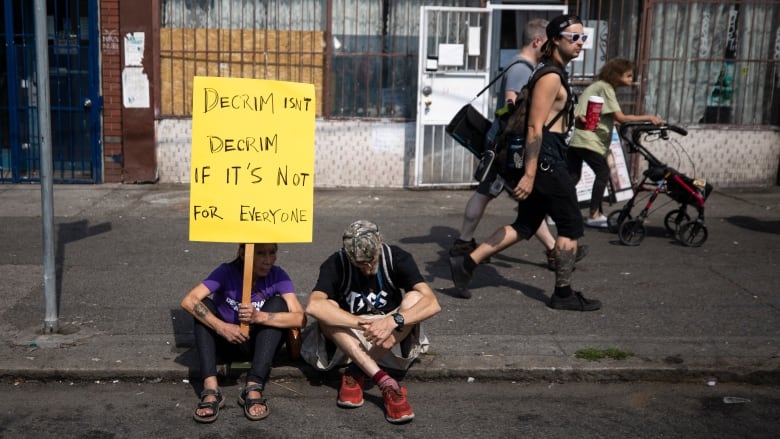 A woman in sandals and a man in red shoes sit on the curb of Vancouver's East Hastings street. The woman has a tatoo on her right arm and is holding a yellow cardboard sign attached to a piece of wood that reads, 