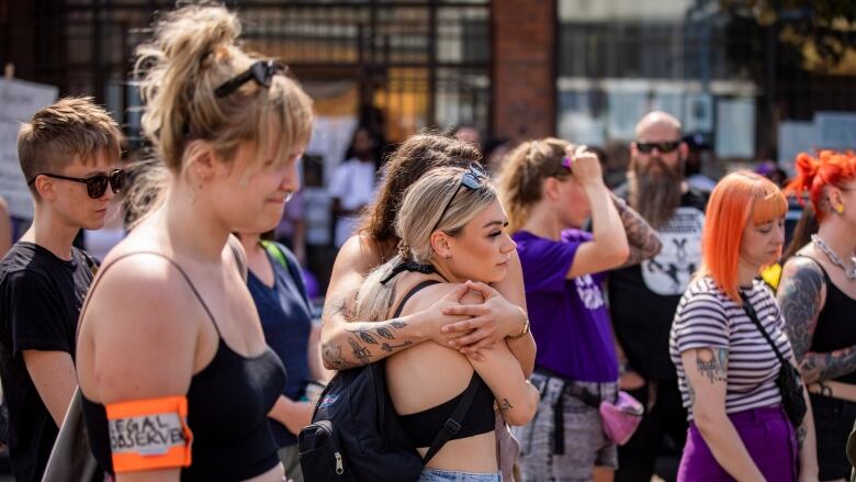 People in a crowd close their eyes, two people embrace and another grimaces as they remember people who have died due to the toxic drug supply at a rally on East Hastings Street in Vancouver, B.C.