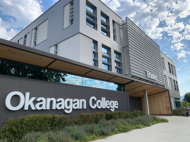 A low-slung, glass-walled building with the sign, 'Okanagan College' connected to a taller grey structure flanked by windows with the world Trades above the entrance.