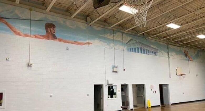 A wide shot of a school gym wall, mostly painted white. The top of a mural depicting a shirtless man throwing a javelin, some clouds, and a building can be seen.