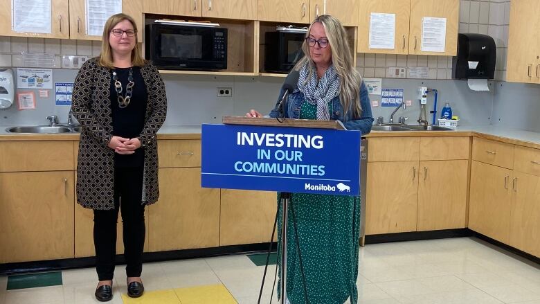 A woman stands speaking at a podium in a classroom kitchen.