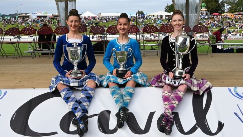 Three girls sit on a stage wearing kilts and matching dancing costumes. They are all carrying trophies. Beneath them, a sign reads 'Cowal Gathering'.