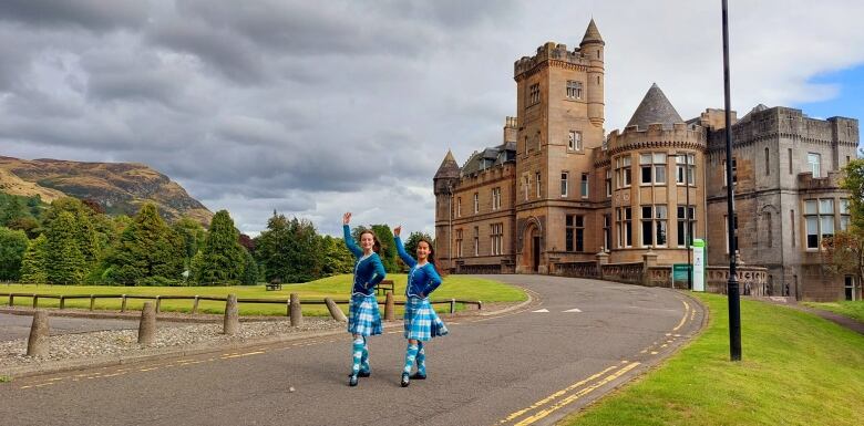 Two girls in identical blue kilts and dance costumes stick their hands up in front of a castle.