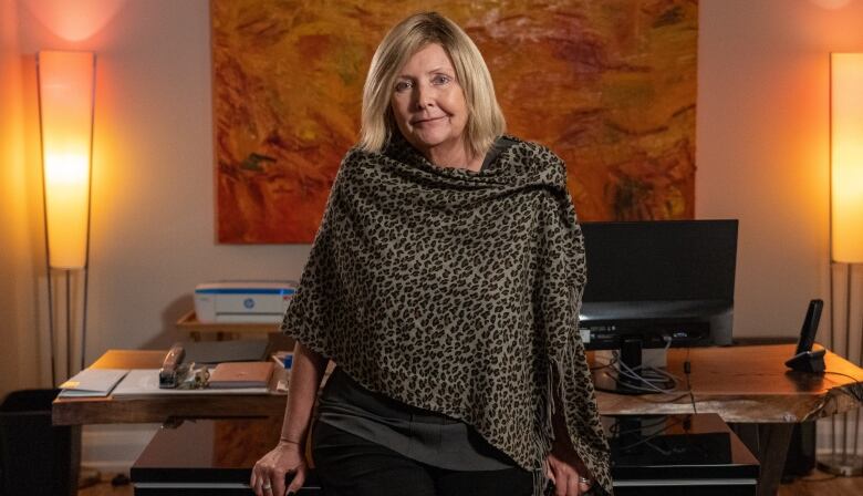 A woman in a shawl leans back against a desk in a personal office