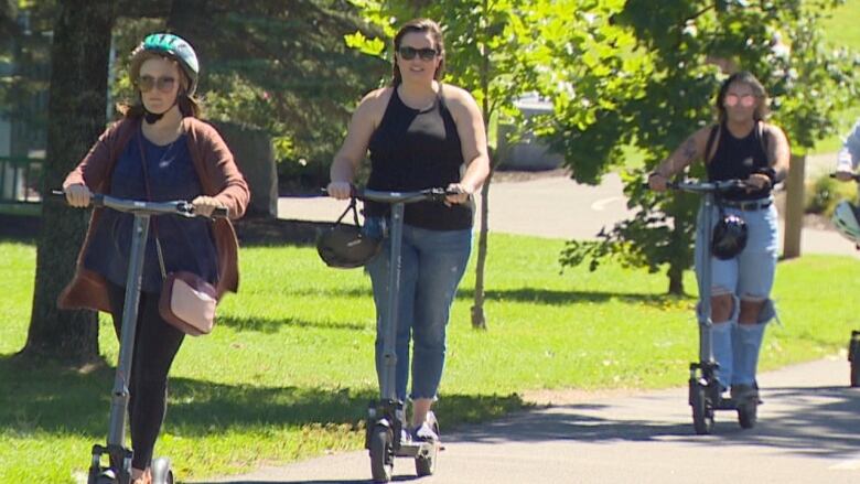 A row of people riding electric scooters move through a park setting. It is summer. 