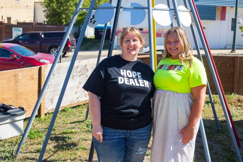 Two women stand in front of a metal tipi with coloured poles.