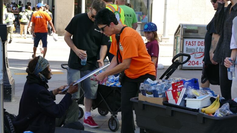 A person in an orange shirt hands an item to a person sitting on a city sidewalk.