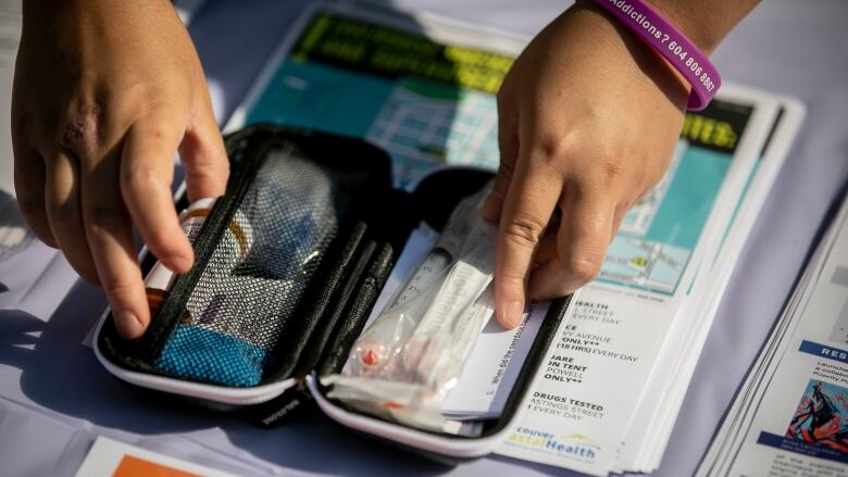 A closeup shows the hands of a person, wearing a purple rubber bracelet, holding a kit containing a syringe and medicine.