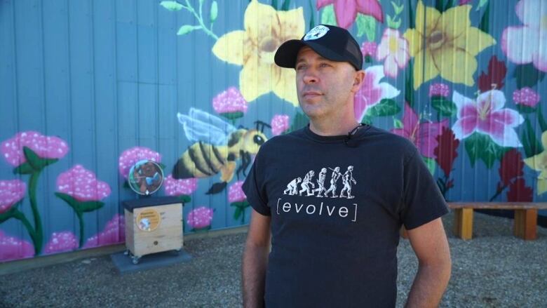 A man in a black shirt stands in front of a mural featuring a bee and flowers.