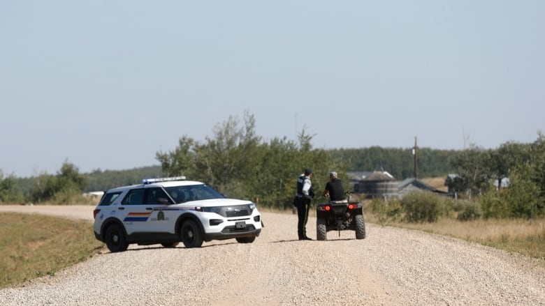 A police officer stands near an RCMP vehicle. The officer is speaking to another person, who is sitting on an ATV.