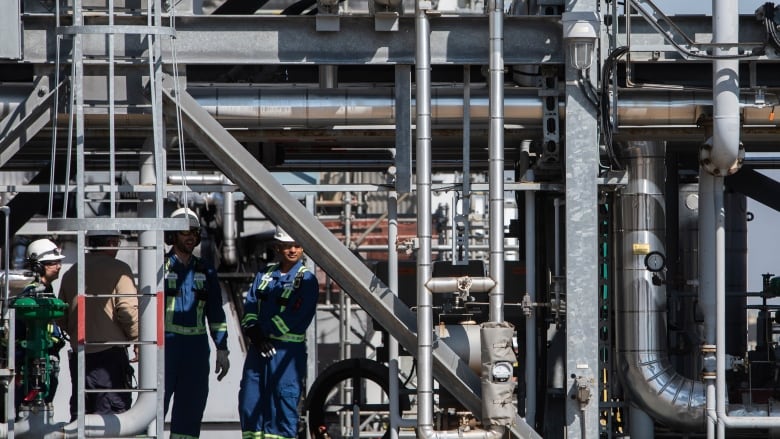 Workers in hard hats and coveralls stand in a hydrogen production plant, amidst pipes and a ladder.