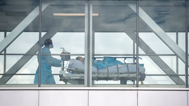 A man in hospital clothing and wearing a face mask pushes a stretcher, with a person lying on it, under a blanket.