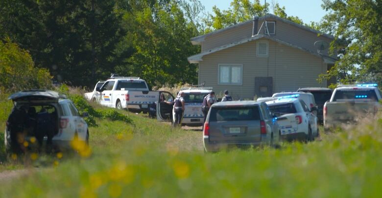 Police vehicles and officers are seen outside a house in a wooded area. 