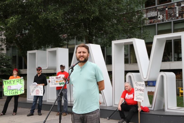 A person speaks at a mic outside before a sign spelling 