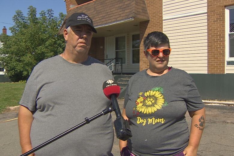 Man and woman stand in front of hotel, looking sad.