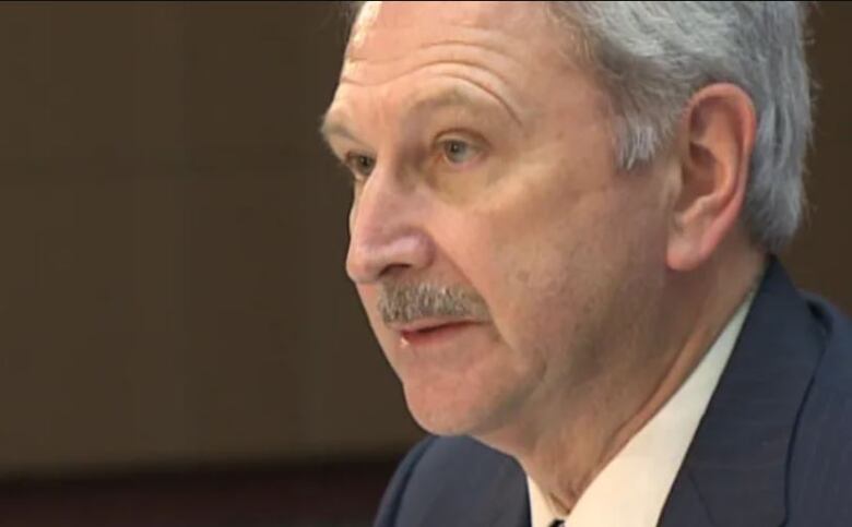 A man with short grey hair and a short grey moustache looks off into the distance to the left of the photo. He's wearing a navy suit over a white dress shirt and dark tie.