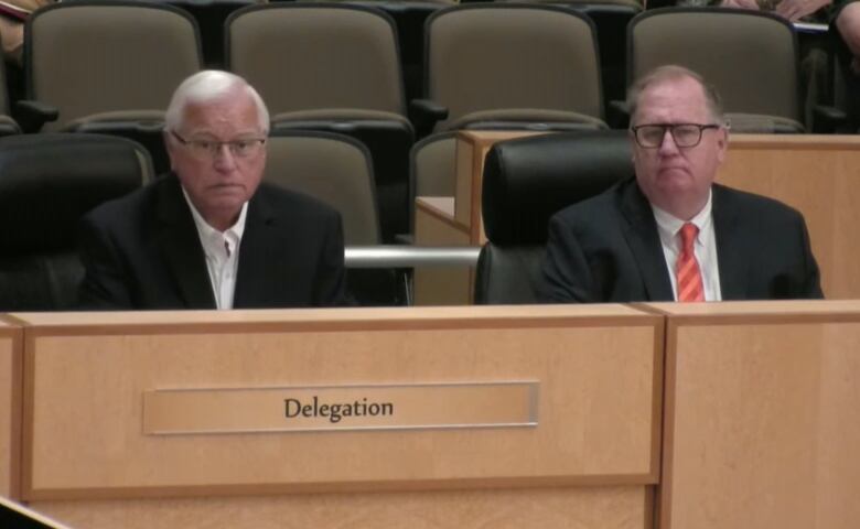 Two men in dark suits sit in chairs in a council chamber in an area marked 