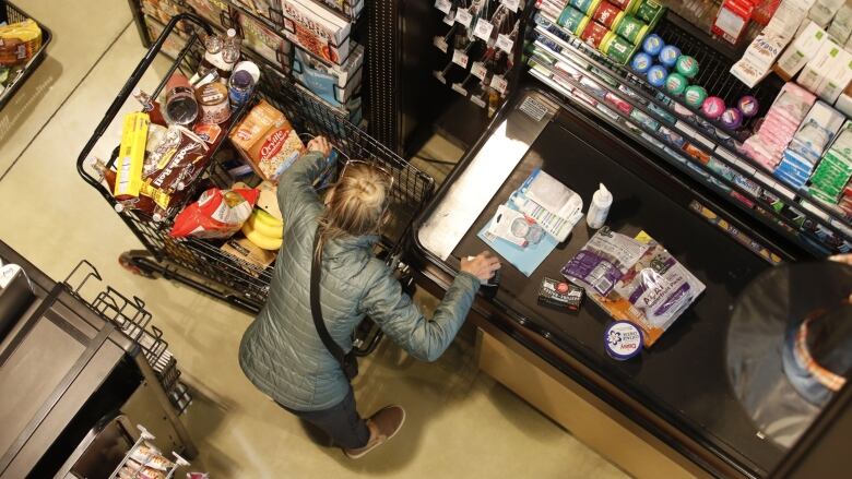 A person, seen from above, unloads items from a full grocery cart onto a conveyor belt at a cash register. 