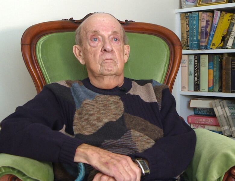 A man, seen from the torso up, sits on a green chair. A book shelf is behind him on the right side.