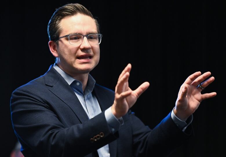 Pierre Poilievre, in a navy blue business jacket and unbuttoned blue shirt, raises his hands to make a point. 