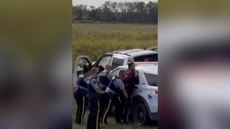 A blurry cellphone photo shows multiple police officers standing behind a man who is pressed up against an RCMP vehicle and has his hands behind his back.