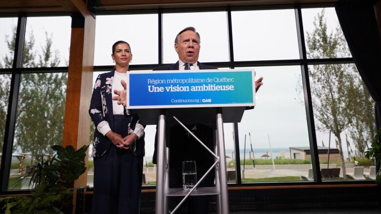 A man stands in front of a stand that's adorned by a campaign sign. A woman stands beside him on the left.