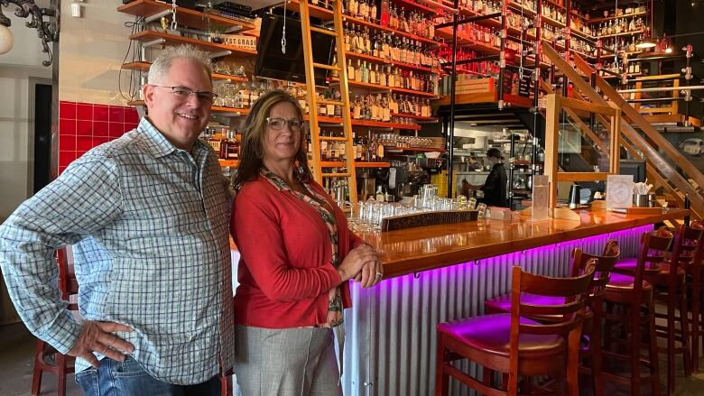 A husband and wife pose in front of a bar, with dozens of bottles behind them. The man is a white man with white spiked hair and glasses. The woman is blonde and is wearing a red top.