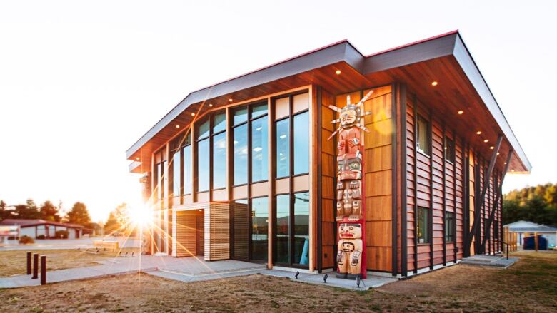 A two-storey wood and glass building, with a totem pole in front, stands in the rising sun.