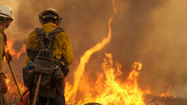 Two men in white helmets and yellow jackets, with bags and tools hanging from their backs, stand in front of flames. 