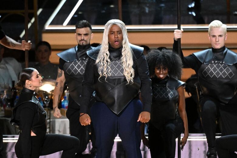 A man wearing a long, blond wig sings on stage, joined by other performers in matching dark leather costumes. 