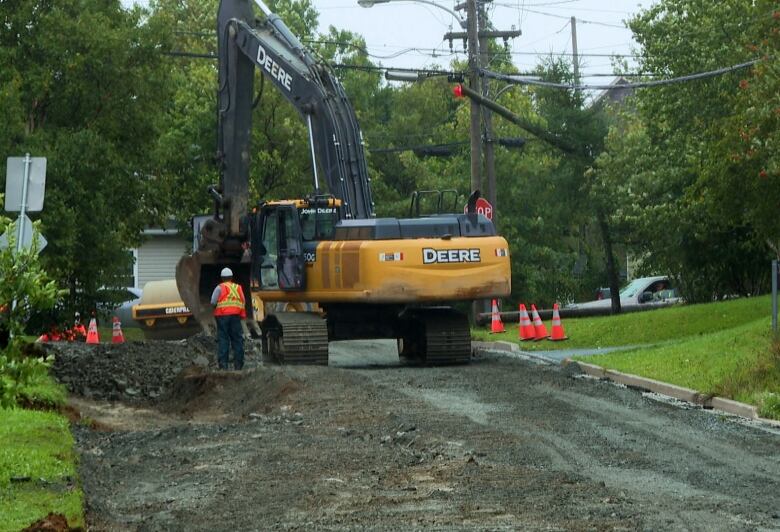 An excavator repairs a road partially washed out on the left side. A person wearing an orange safety vest and hard hat stands near the excavator.