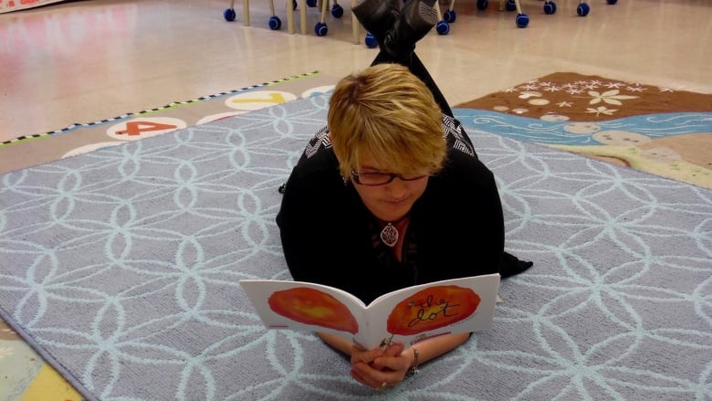 A woman reads a story book lying on her stomach in the middle of a classroom.