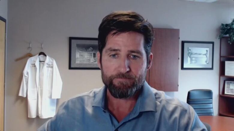 A man in a blue shirt sits in an office, with a white lab coat hanging on the wall behind him.