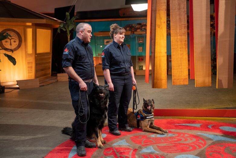 A man and woman wear a black uniform while holding the leashes of two dogs.