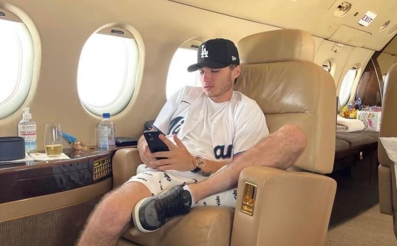 A young man sits in the seat of a private jet looking at his phone. 