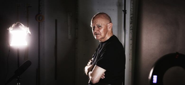 Older bald man standing with arms folded across his chest with a light shining on him.