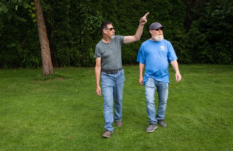 Two men with grey beards are shown walking and talking with each other in a park. One of them is pointing at something in the sky.