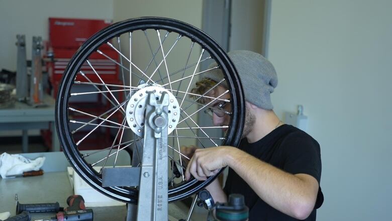 A man wearing a t-shirt and toque works on a wheel locked into a vice. 