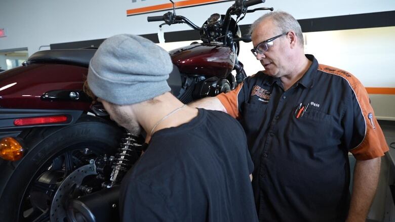 In a garage bay, a student looks at a motorcycle exhaust pope that an instructor points at. 