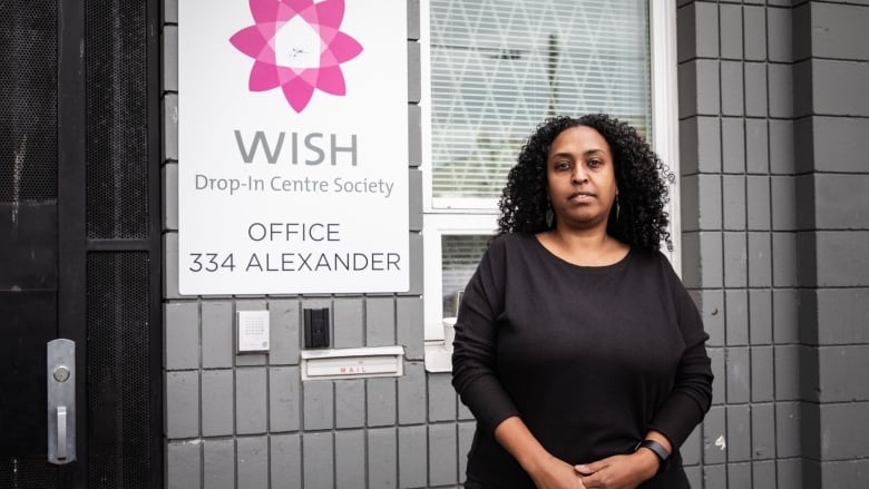 A Black woman stands in front of a sign that says 'WISH Drop-In Centre Society'.