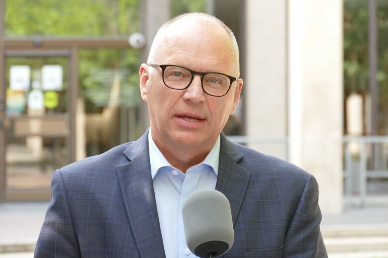 A man wearing glasses and a suit is speaking into a microphone, with a building in the background.