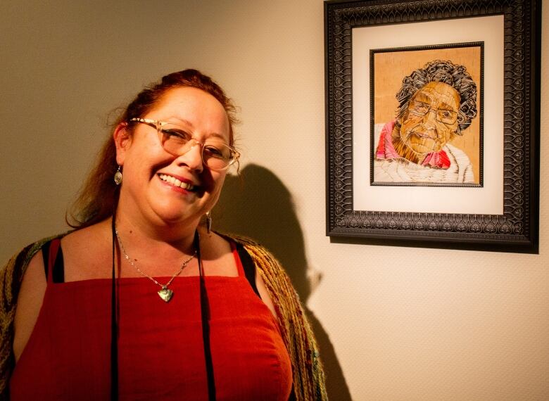 Woman in red dress and glasses smiles beside an artwork depicting an elderly woman, entirely made out of porcupine quills.