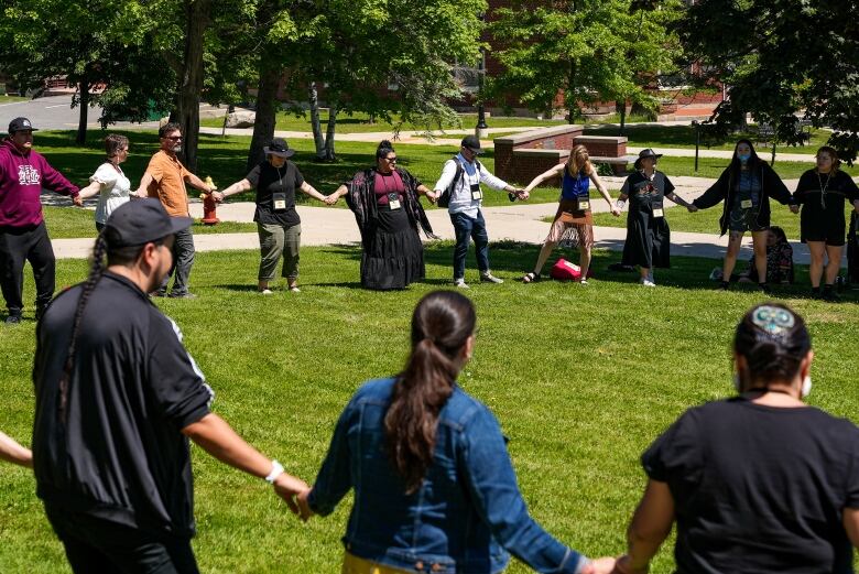 People dance, holding hands, in a circle on a grassy lawn.