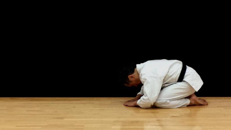 Man wearing a white karate uniform in a deep bow on a polished wood floor against a black background.